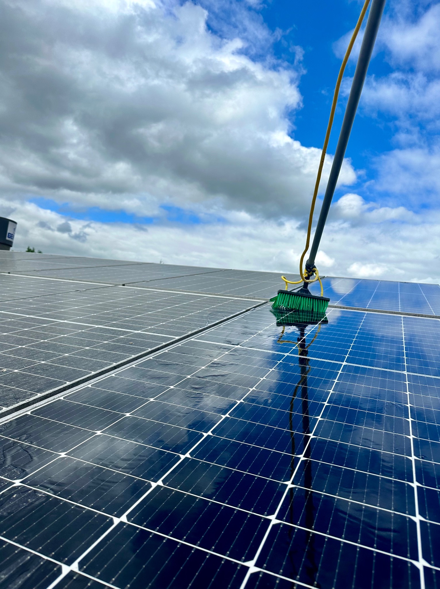 Szczotka na długim wysięgniku czyści panele słoneczne, odbicie nieba w mokrej powierzchni paneli.