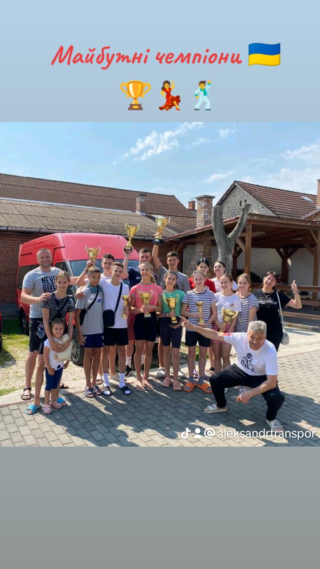 Grupa dzieci i dorosłych trzyma złote puchary przed czerwonym busem, napis 'Majbutni Czempiioni' i flaga Ukrainy u góry