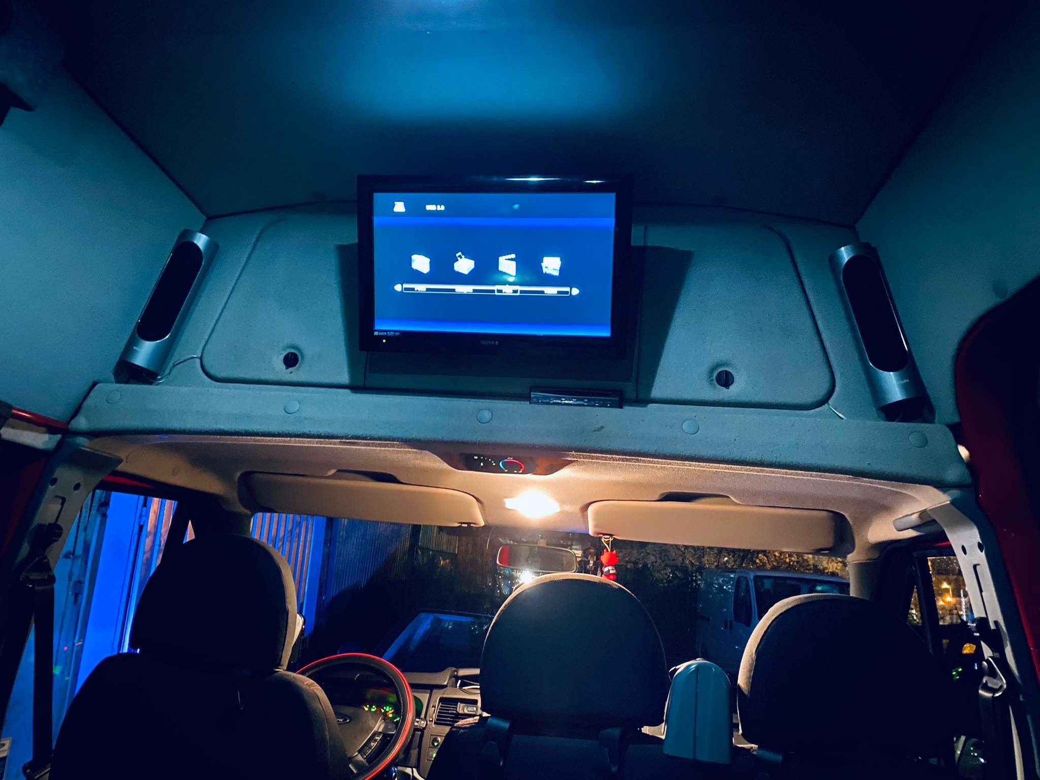 Wnętrze busa z zamontowanym telewizorem dla pasażerów, widoczne fotele i deska rozdzielcza kierowcy, oświetlenie wnętrza.