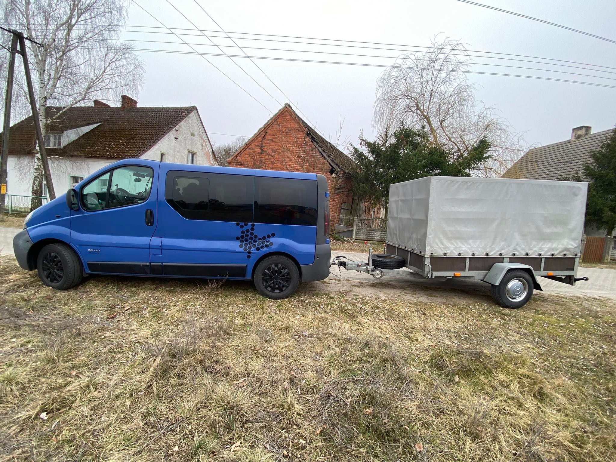 Niebieski bus z czarnymi felgami i naklejką w kształcie plastra miodu ciągnie przyczepę z plandeką na tle wiejskich zabudowań i pochmurnego nieba.