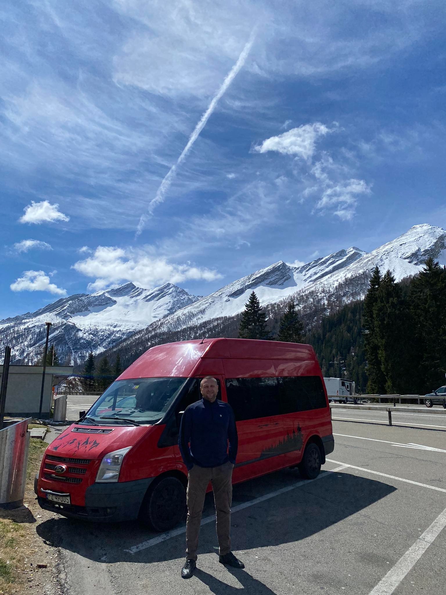 Czerwony bus Ford Transit z grafiką lasu na boku, zaparkowany na tle górskiego krajobrazu z ośnieżonymi szczytami i błękitnym niebem z chmurami.