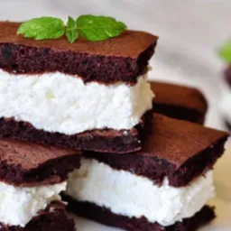 Stos brązowych kanapek z ciasta z kremowym, białym nadzieniem, udekorowanych listkami mięty na jasnym tle.