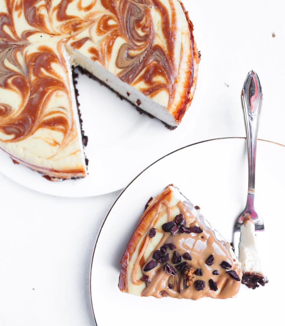 Sernik z wirującymi wzorami karmelu na białym tle, obok talerzyk z kawałkiem ciasta polanym sosem i posypanym kawałkami czekolady, elegancki widelec.