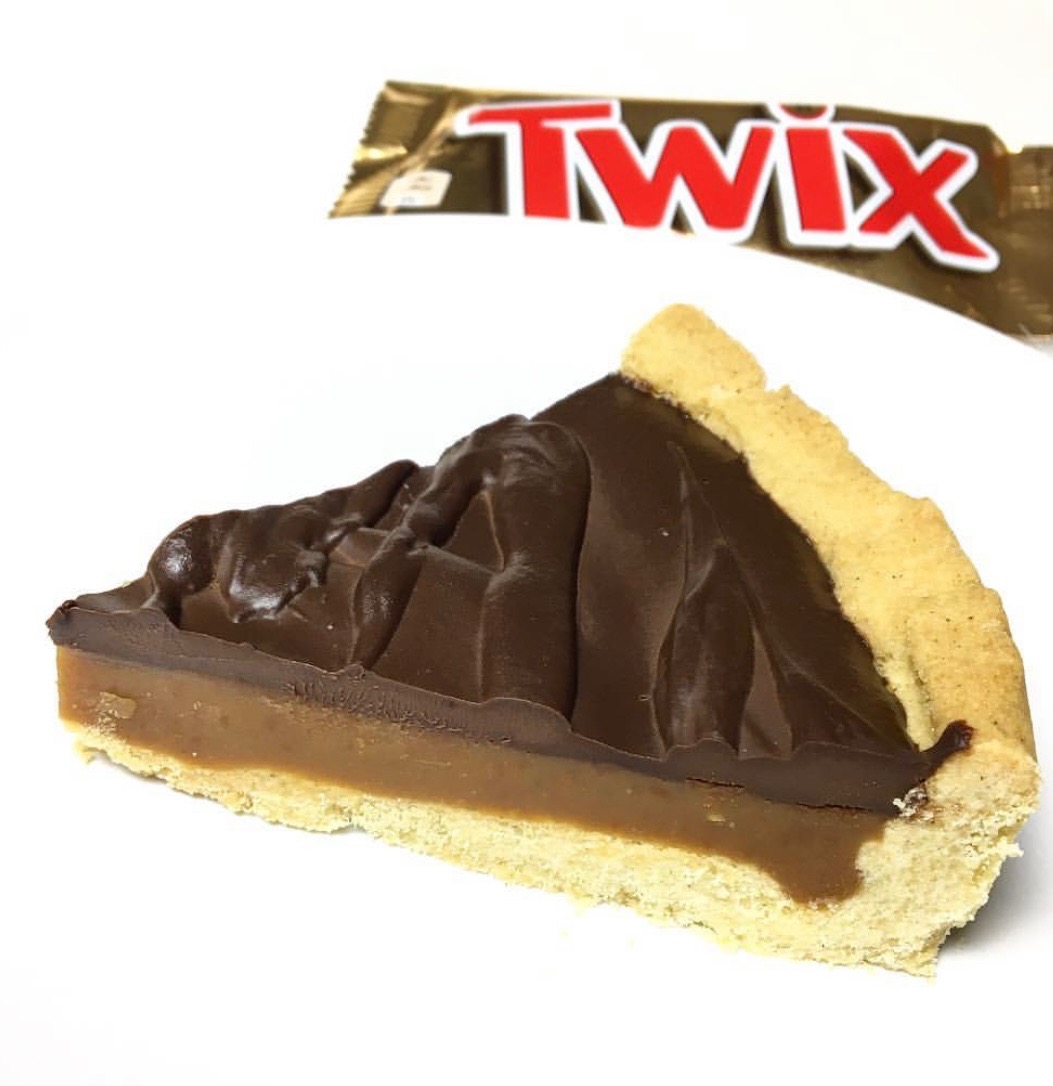 Kawałek tarty Twix z kruchym spodem, warstwą karmelu i polewą z mlecznej czekolady, na białym tle z opakowaniem batonika Twix w tle.