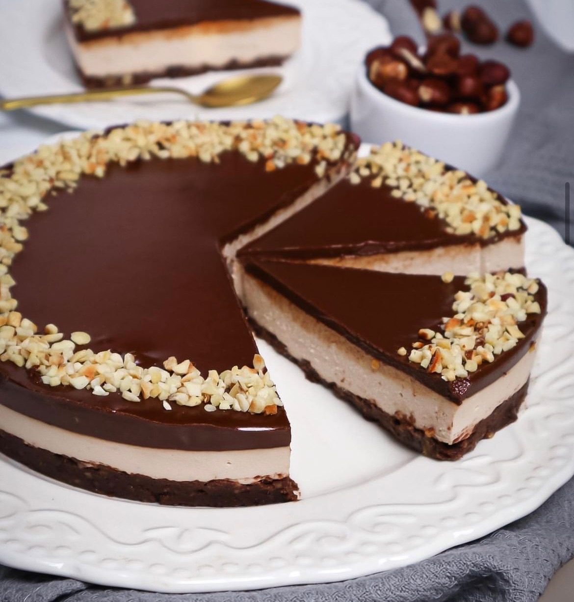 Tort z czekoladową polewą i posiekanymi orzechami laskowymi, z widocznym przekrojem ukazującym warstwy kremu, na białym talerzu z dekoracyjnym brzegiem, w tle miseczka z orzechami i kawałek tortu.