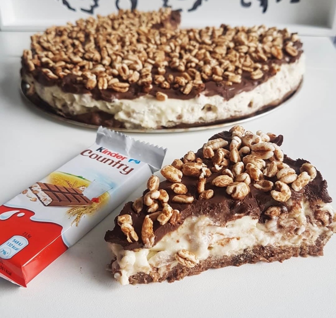Kawałek tortu z kremem i polewą czekoladową, posypany prażonym ryżem, obok batonik Kinder Country.
