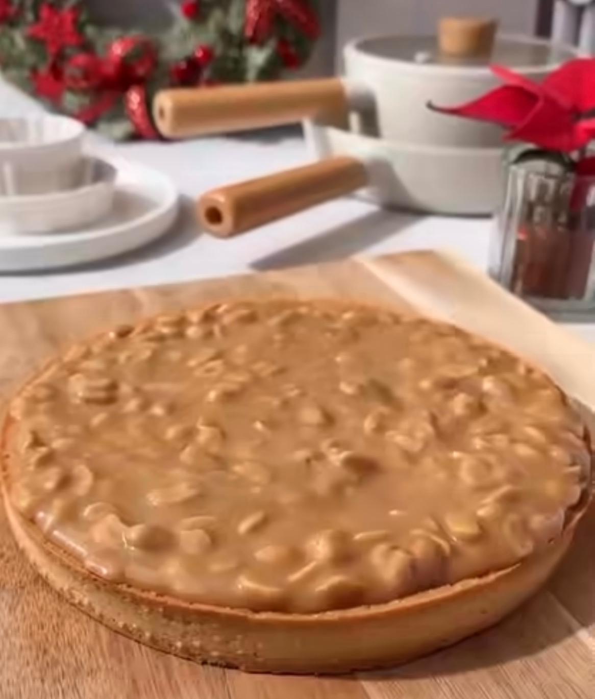 Tarta serowa z polewą karmelową i orzechami na drewnianej desce, w tle świąteczny wieniec i naczynia.