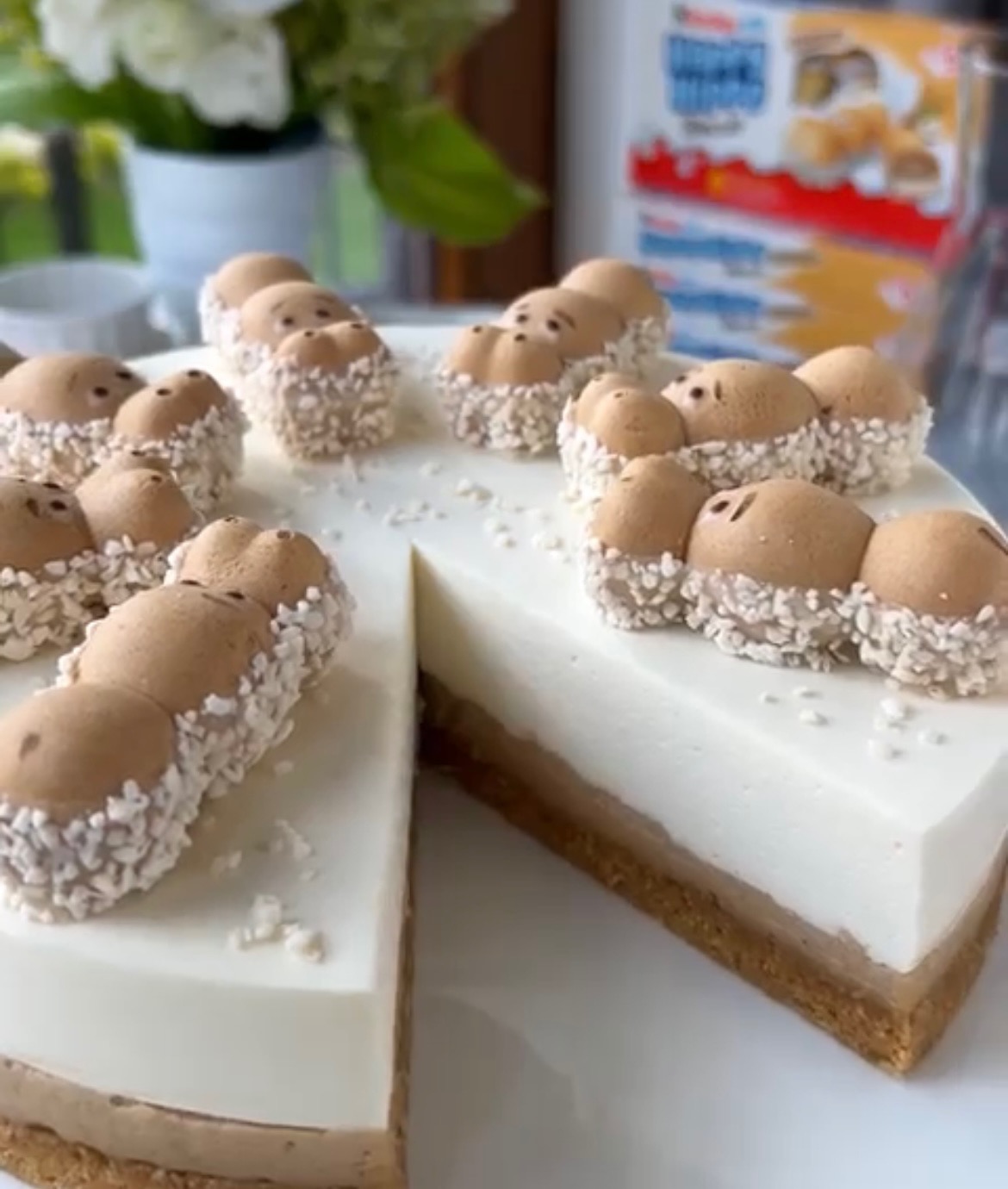 Kawałek tortu z trzema warstwami (biszkopt, krem czekoladowy, krem biały) udekorowany figurkami Kinder Hippo obtoczonymi w białej posypce, na tle pudełka Kinder Hippo i kwiatów.