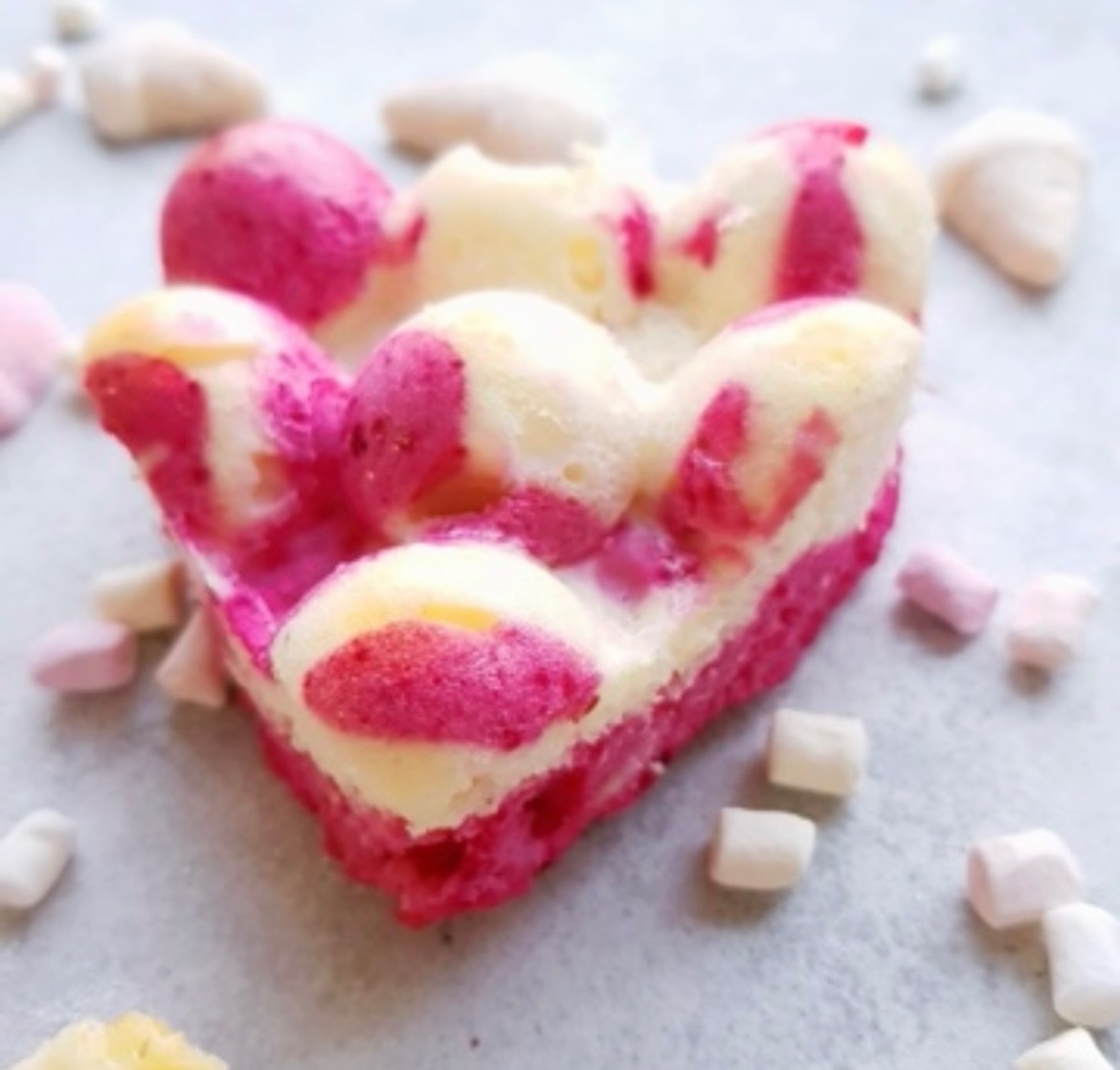 Biszkoptowe serce z różowo-białymi smugami, otoczone mini piankami na szarym tle.