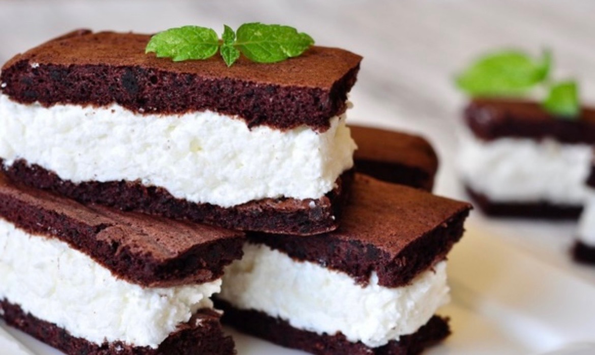 Stos brązowych kanapek z ciasta z kremowym, białym nadzieniem, udekorowanych listkami mięty na jasnym tle.