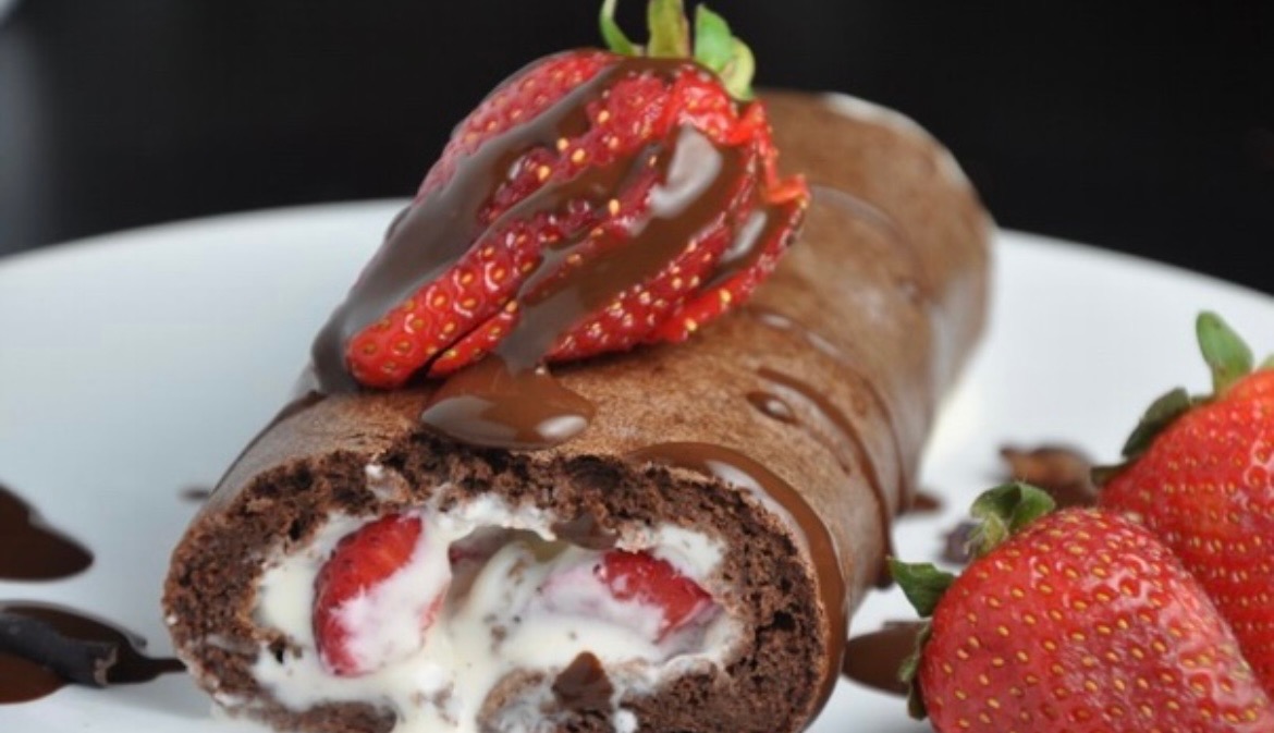 Czekoladowa rolada z kremem śmietankowym i truskawkami, polana polewą czekoladową, udekorowana świeżymi truskawkami na białym talerzu.