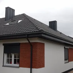 Dom jednorodzinny z ciemną dachówką, oknami dachowymi i ciemnymi rynnami, wykończony cegłą klinkierową na narożnikach i z roletami zewnętrznymi.