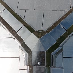 Usługi Ciesielsko Dekarskie - Szczegółowe ujęcie czarnego dachu z ceramicznej dachówki z widocznym systemem odprowadzania wody w kształcie litery Y i fragmentem buta dekarza.
