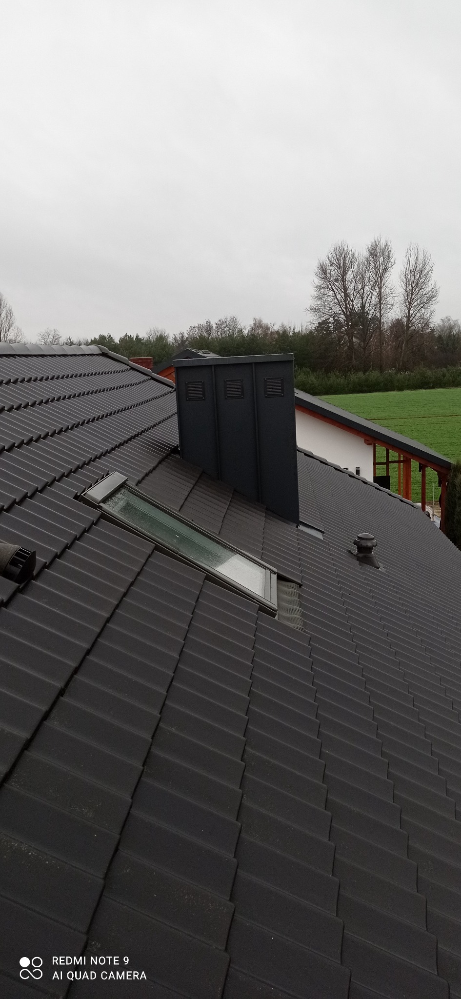 Czarny dach z dachówki ceramicznej z oknem dachowym i kominem wentylacyjnym, widoczne drzewa w tle.