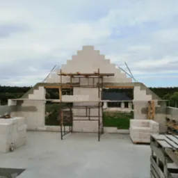 Wznoszenie szczytu domu z białych bloczków, widoczne rusztowanie, wystające pręty zbrojeniowe i palety z materiałem budowlanym na tle zieleni i zabudowań.