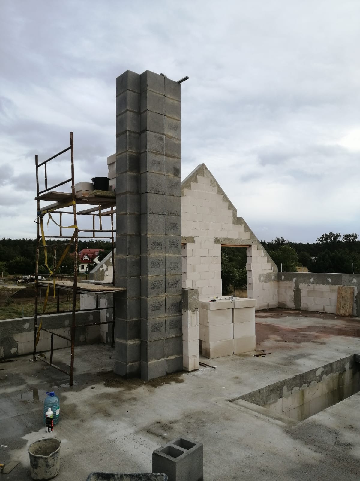Wznoszenie komina z szarych bloczków betonowych przy ścianie domu w trakcie budowy, widoczne rusztowanie i materiały budowlane na placu budowy.
