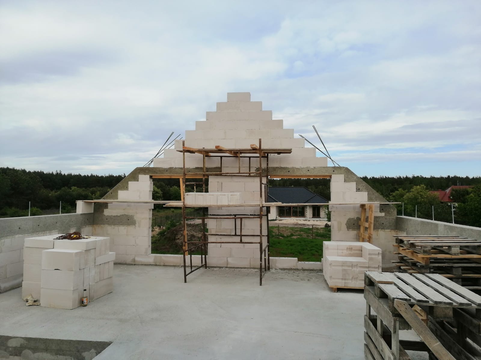 Wznoszenie szczytu domu z białych bloczków, widoczne rusztowanie, wystające pręty zbrojeniowe i palety z materiałem budowlanym na tle zieleni i zabudowań.