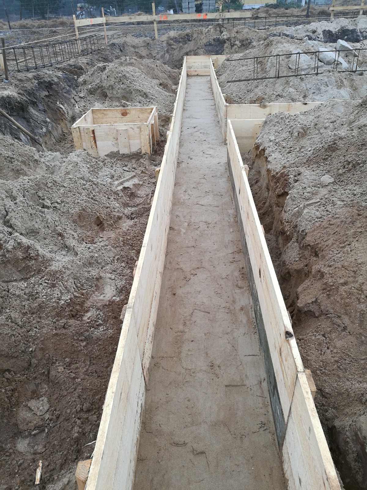 Szczegółowy widok wykopu z drewnianym szalunkiem przygotowanym pod wylewkę fundamentów, widoczne zbrojenia i ziemia.