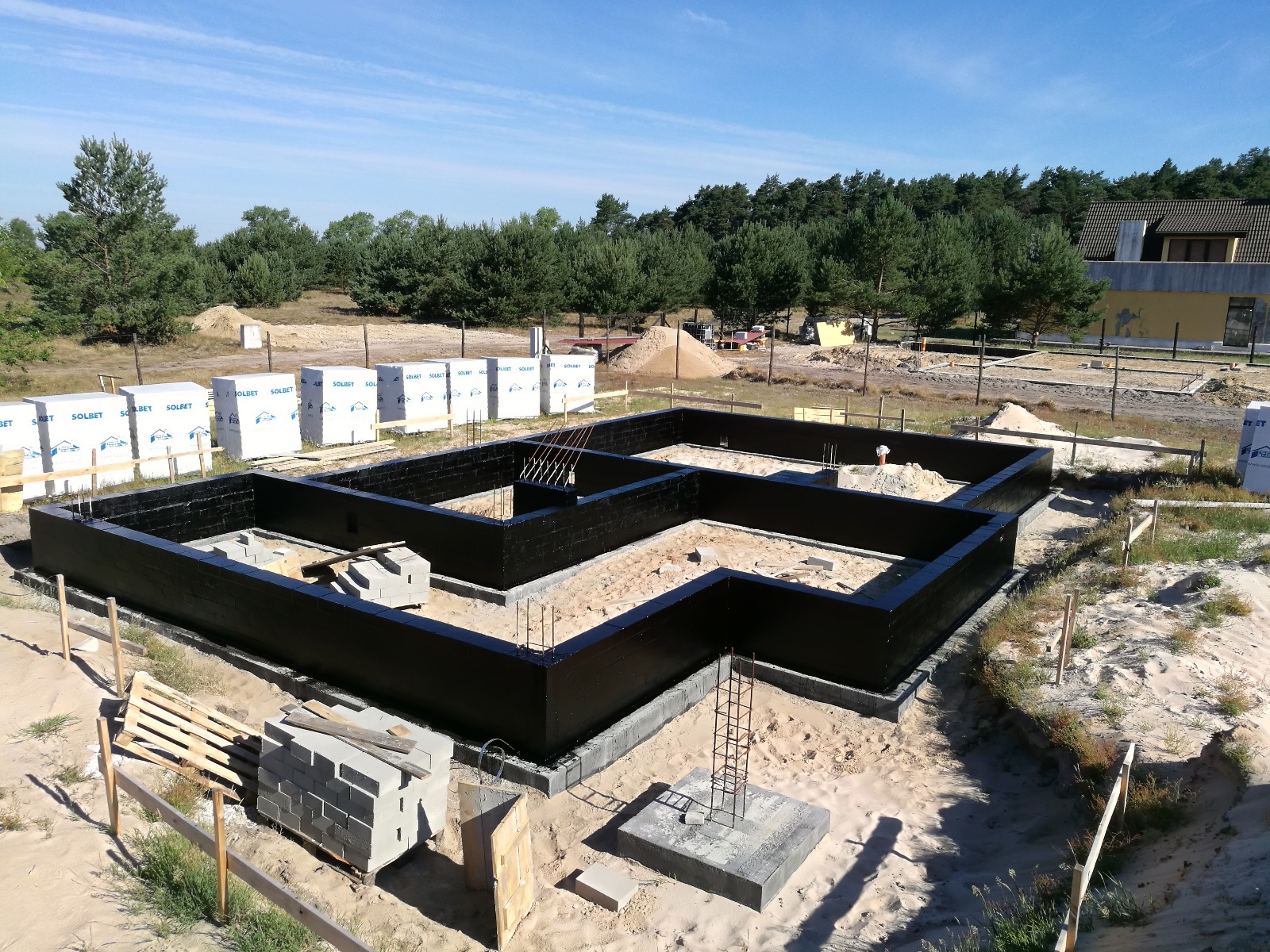 Zalane fundamenty domu jednorodzinnego z hydroizolacją, widoczne zbrojenia, bloczki Solbet i piasek na działce budowlanej w słoneczny dzień.