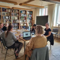 Grupa osób siedzi przy stole podczas spotkania w sali z biblioteczką, laptop i projektor na stole.