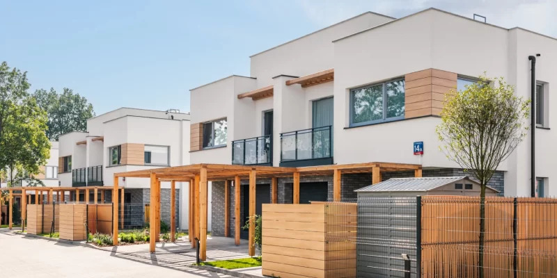 Nowoczesne, białe domy szeregowe z drewnianymi elementami dekoracyjnymi elewacji i pergolami, widok z zewnątrz w słoneczny dzień.