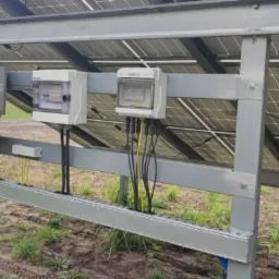 Instalacja fotowoltaiczna: widok od spodu na panele, inwerter i skrzynki elektryczne zamontowane na metalowej konstrukcji na zewnątrz budynku.