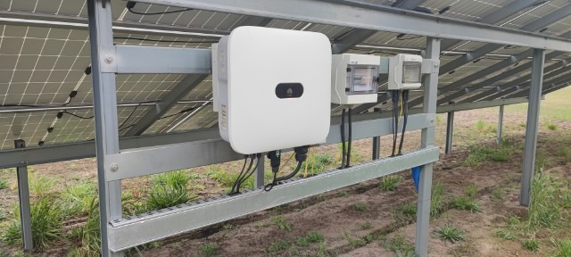 Biały falownik z logo Huawei zamontowany na metalowej konstrukcji pod panelami słonecznymi, wraz z dwiema szarymi skrzynkami przyłączeniowymi i okablowaniem.