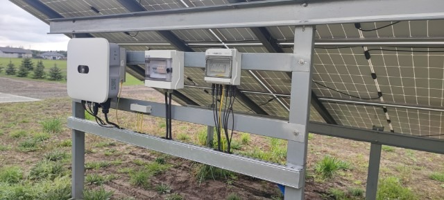 Instalacja fotowoltaiczna: widok od spodu na panele, inwerter i skrzynki elektryczne zamontowane na metalowej konstrukcji na zewnątrz budynku.