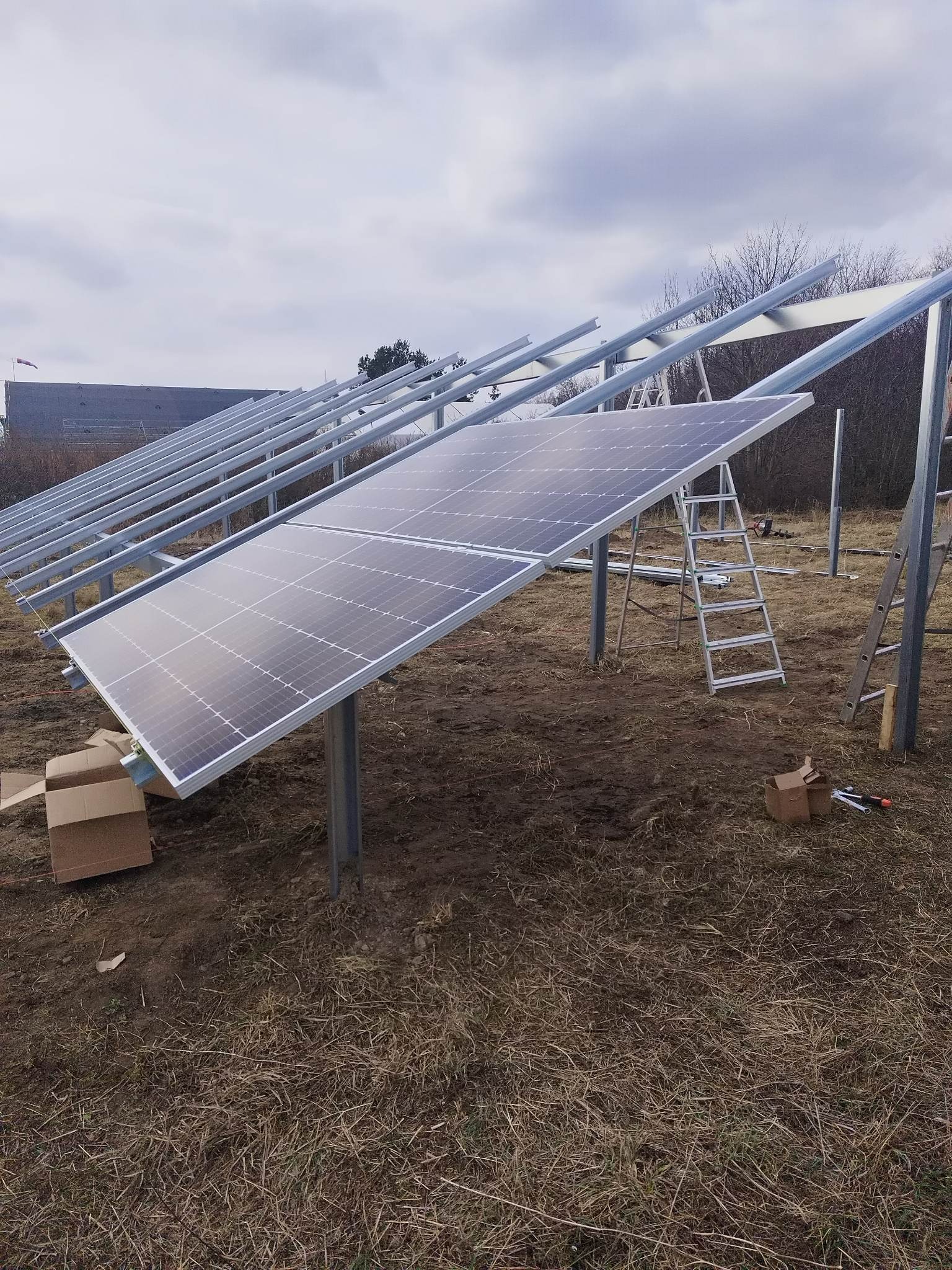 Montaż paneli słonecznych na metalowej konstrukcji, widoczne drabiny i narzędzia na zewnątrz w pochmurny dzień.