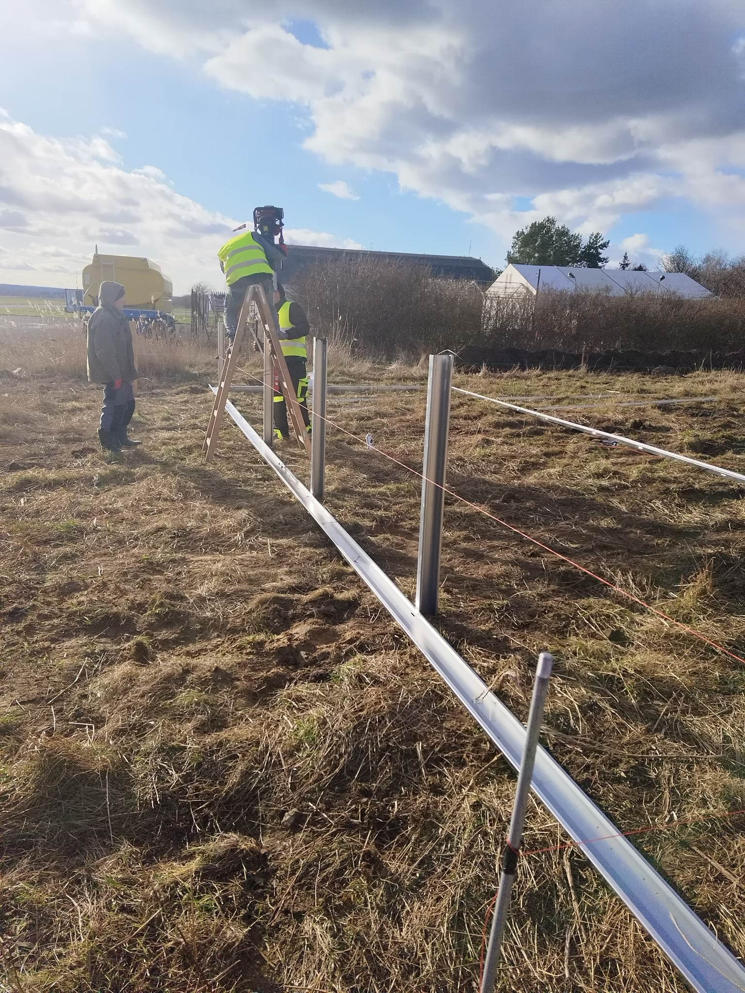 Montaż konstrukcji wsporczej na polu: dwóch pracowników w kamizelkach na drabinie ustawia metalowe słupki i szyny, trzeci obserwuje, w tle budynek gospodarczy i pochmurne niebo.