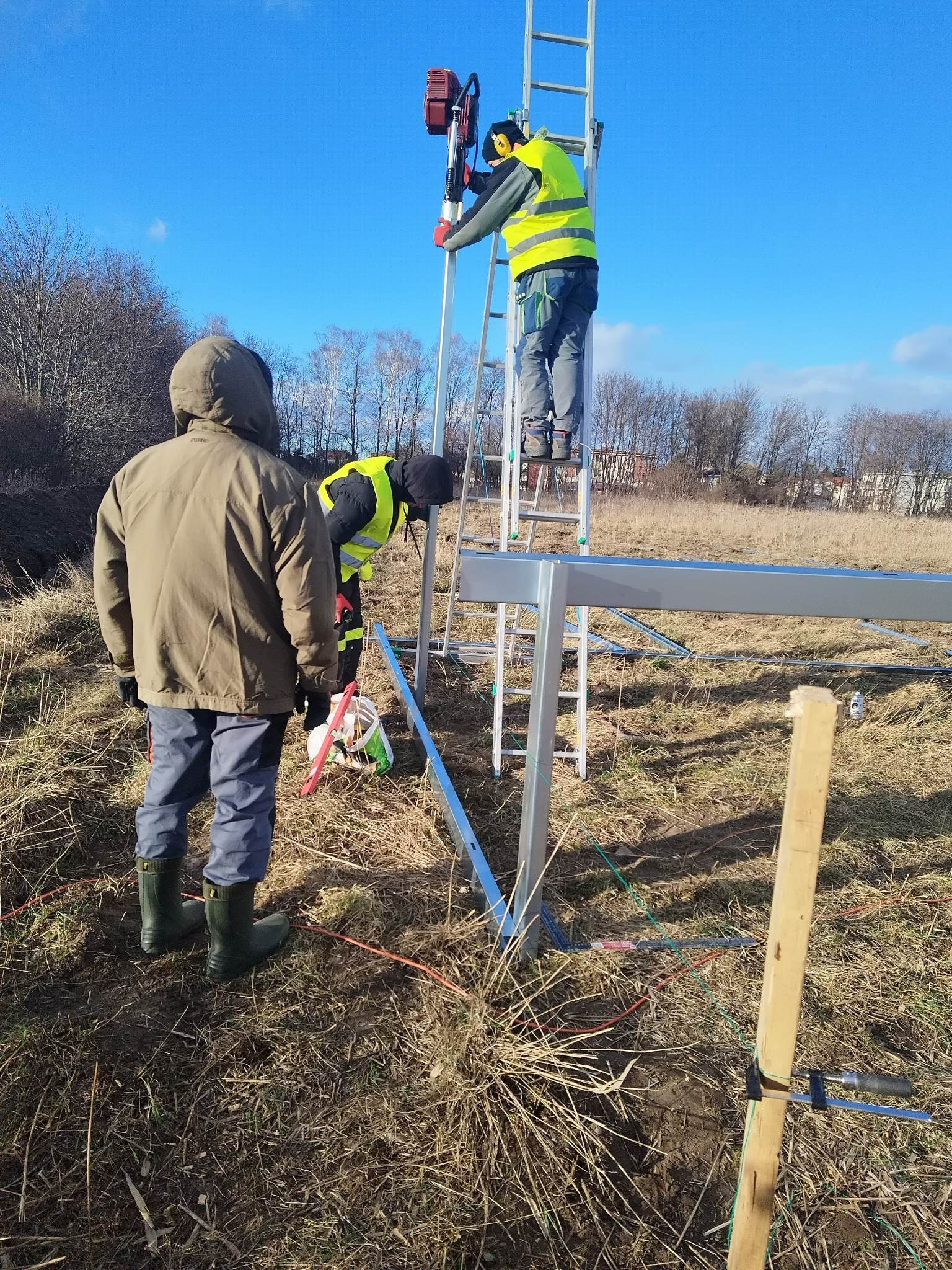 Dwóch monterów w odblaskowych kamizelkach instaluje konstrukcję na otwartym terenie; jeden na drabinie używa narzędzia, drugi stoi obok, w tle błękitne niebo i drzewa.