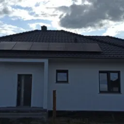 Nowoczesny dom jednorodzinny z ciemną dachówką i panelami słonecznymi na dachu, widok z poziomu gruntu, błękitne niebo z chmurami w tle.