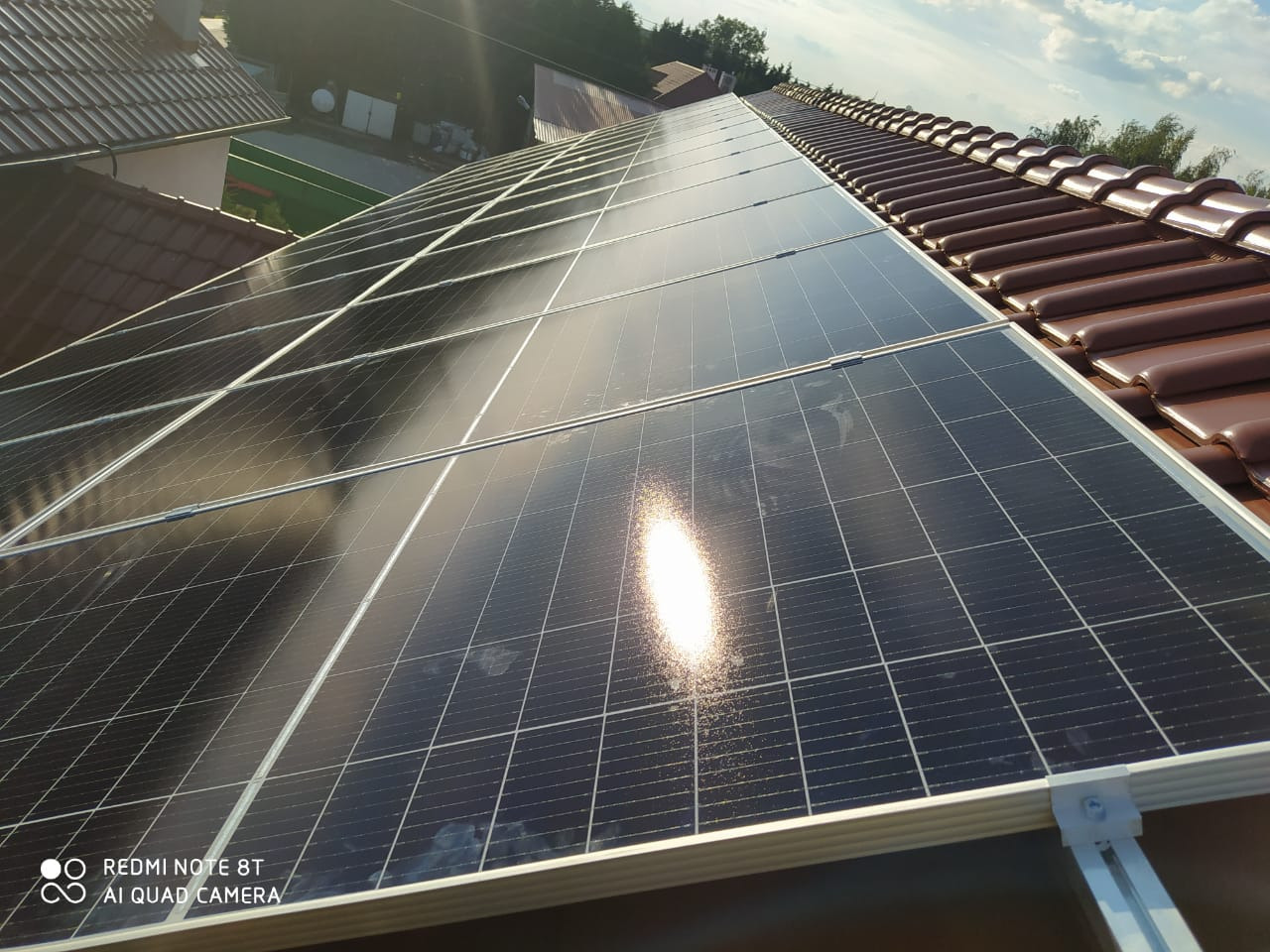 Panele fotowoltaiczne zamontowane na brązowym dachu z dachówki ceramicznej, widoczne elementy montażowe i odbicie słońca na powierzchni paneli.