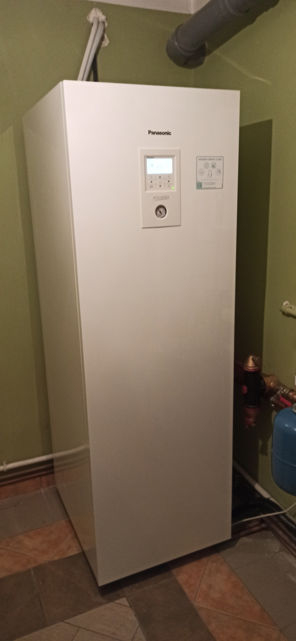 Biała pompa ciepła Panasonic Aquarea zainstalowana w pomieszczeniu z zielonymi ścianami i brązową podłogą, widoczne rury i przewody doprowadzające.