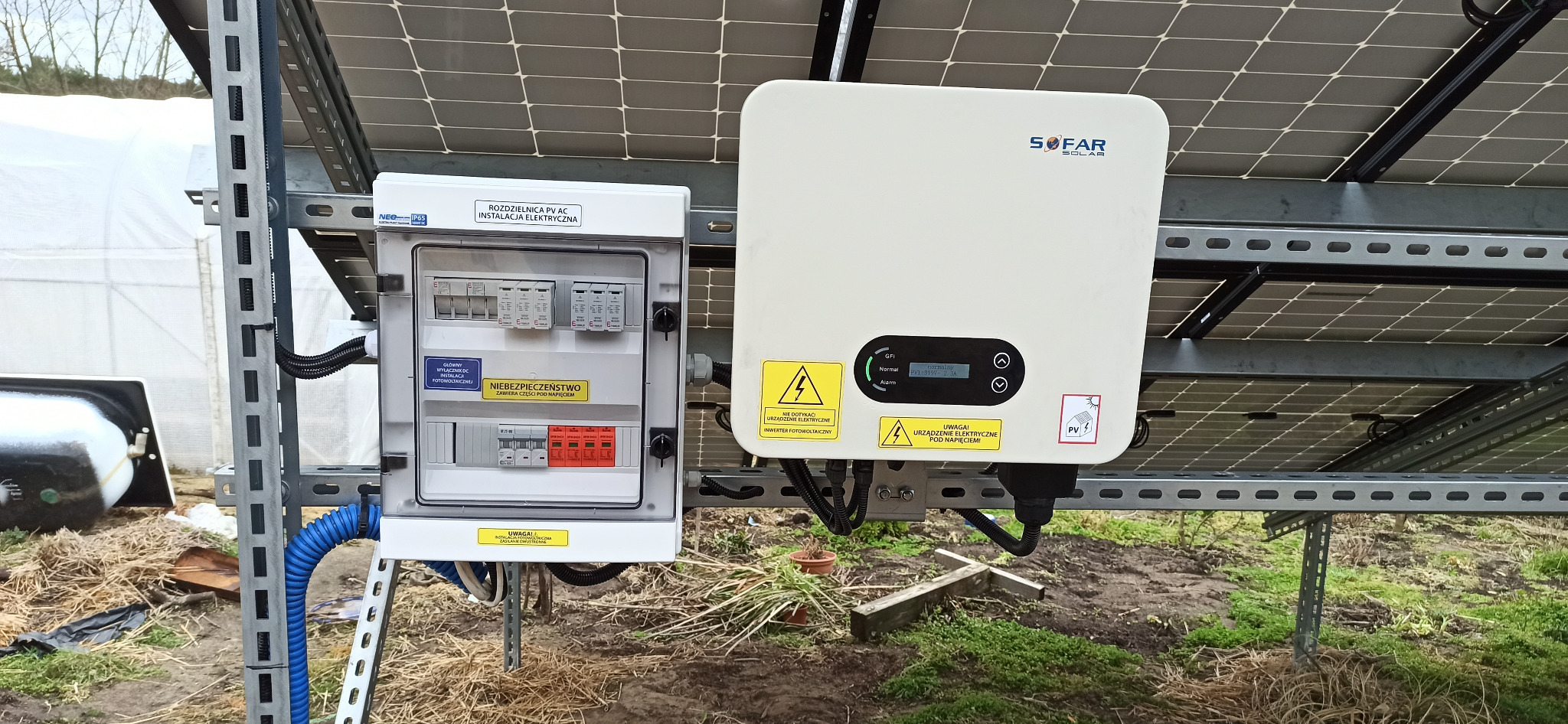 Rozdzielnica AC/DC i inwerter SOFAR Solar przy panelach fotowoltaicznych, widoczne zabezpieczenia i okablowanie, instalacja w terenie.