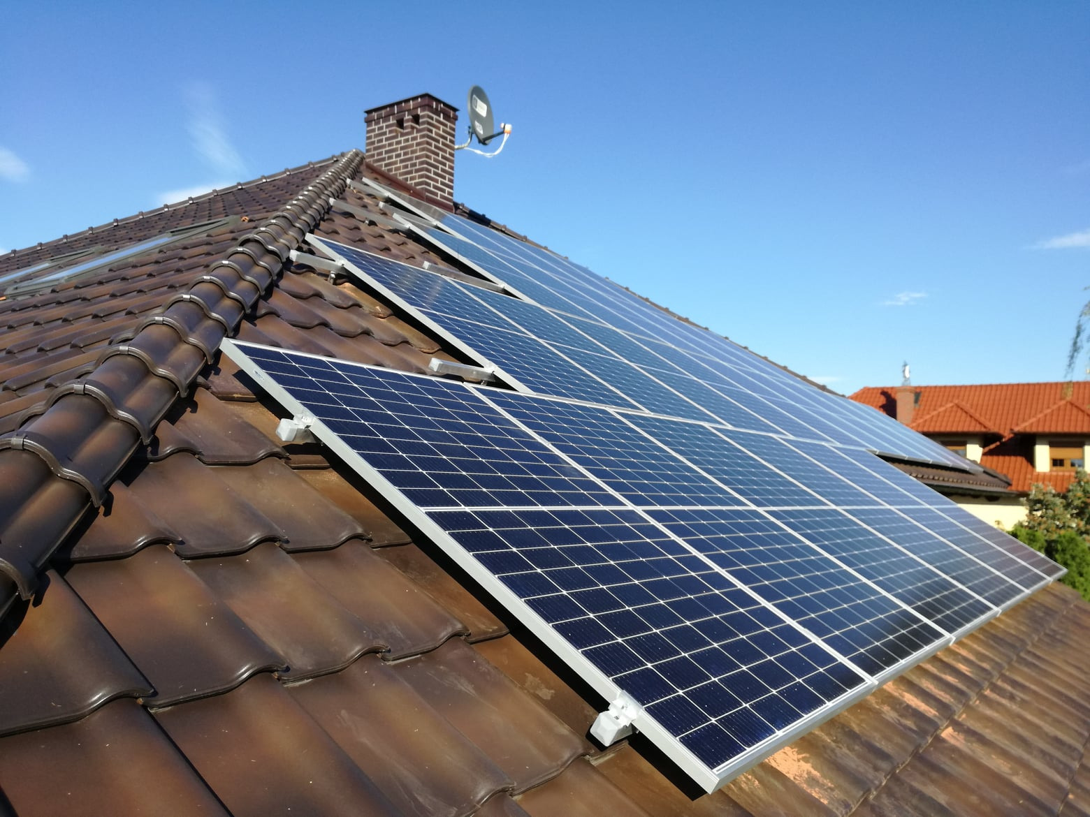Panel słoneczny na brązowym dachu z dachówki ceramicznej, widoczny komin z anteną satelitarną na tle błękitnego nieba.