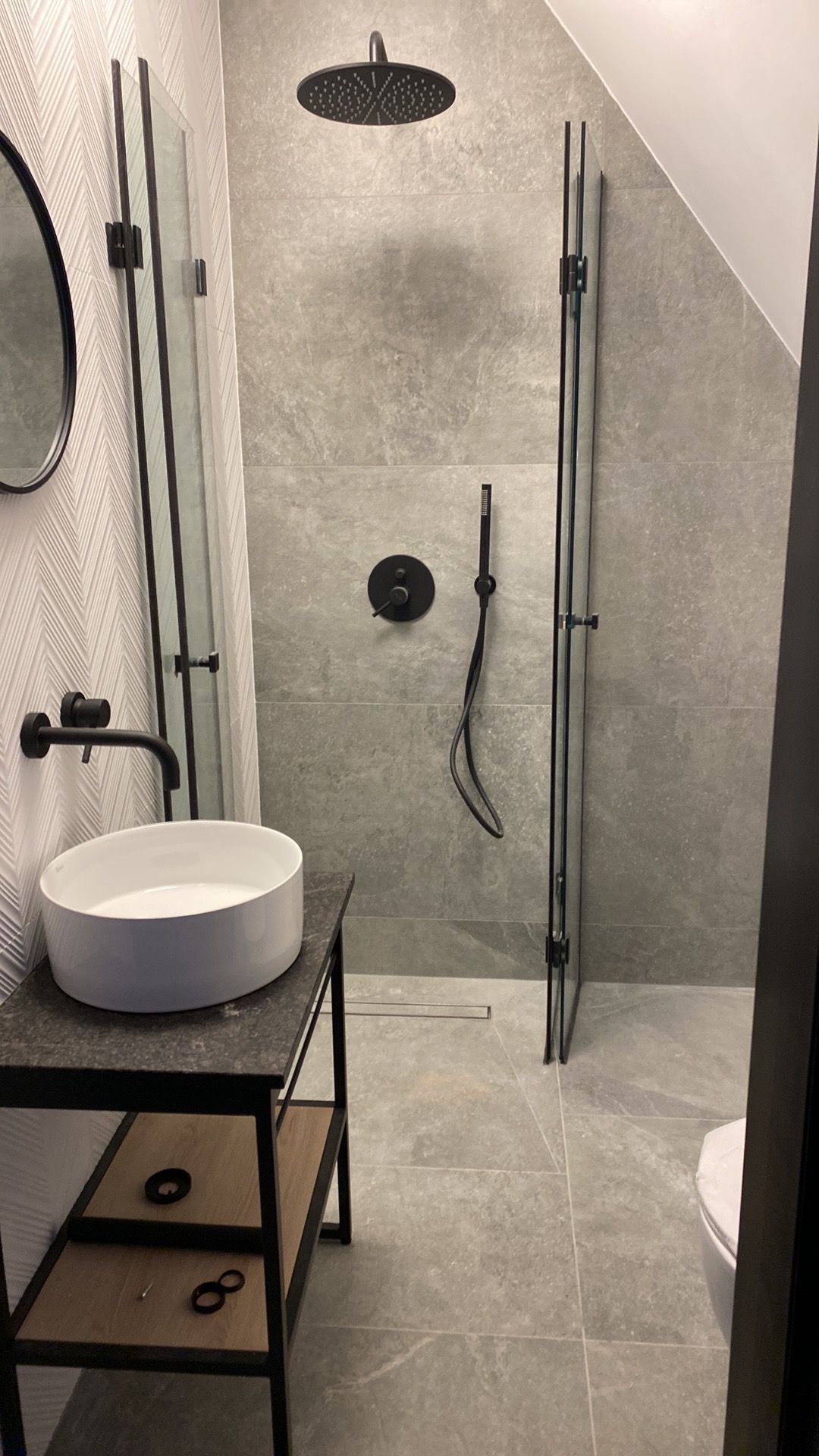 Nowoczesna łazienka w szarościach z czarnymi akcentami: kabina prysznicowa typu walk-in, okrągła umywalka nablatowa, bateria i deszczownica w kolorze czarnym, płytki imitujące kamień, skośny sufit.