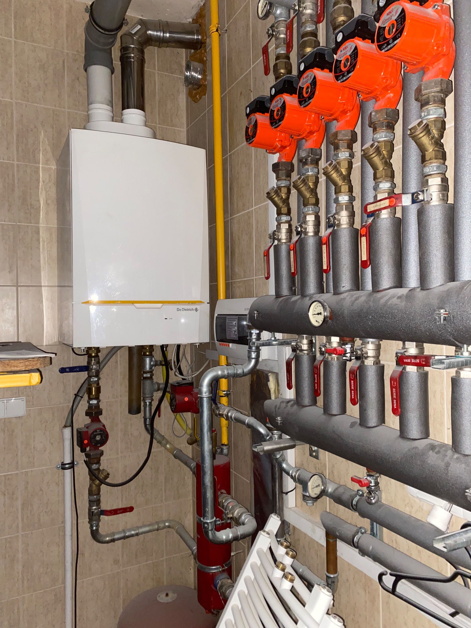 Biały piec gazowy De Dietrich zamontowany na ścianie z beżowych płytek, widoczne rury, zawory, pompy obiegowe w kolorze pomarańczowym i grzejnik panelowy w tle. Instalacja grzewcza z izolowanymi...