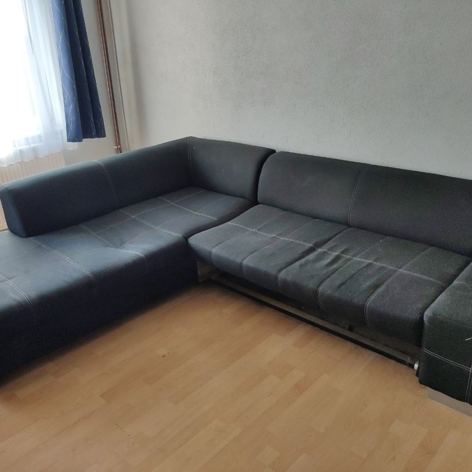 Ciemnoszara narożna kanapa z widocznymi przeszyciami i lekkimi śladami użytkowania, ustawiona na drewnianej podłodze przy ścianie i oknie z ciemną zasłoną.