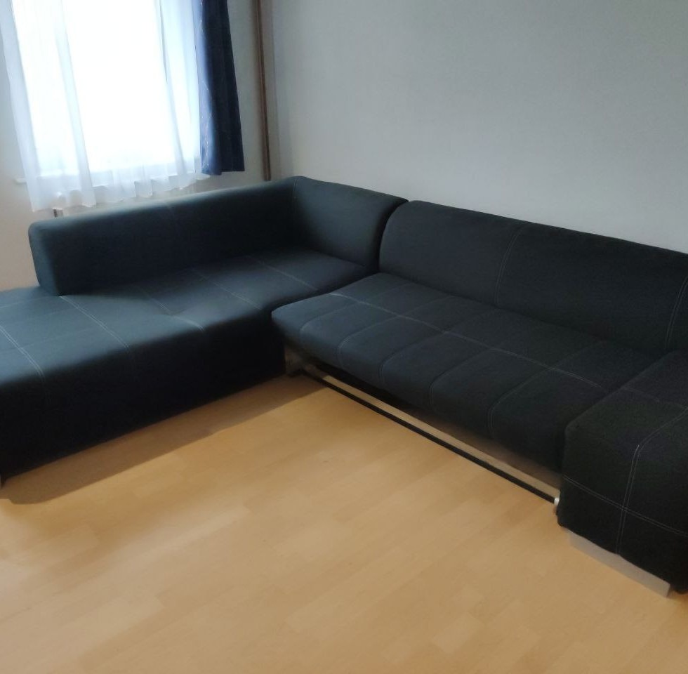 Ciemnoszary narożnik z funkcją spania i jasnymi przeszyciami, ustawiony na parkiecie w pokoju z oknem zasłoniętym granatową zasłoną.