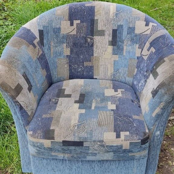 Fotel tapicerowany wzorzystą tkaniną w odcieniach niebieskiego i szarości, z widocznymi elementami trawy w tle.