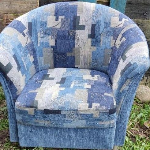 Krótkie krzesło tapicerowane w odcieniach niebieskiego i szarego z geometrycznym wzorem, stojące na zewnątrz na tle drewnianego płotu i zieleni.