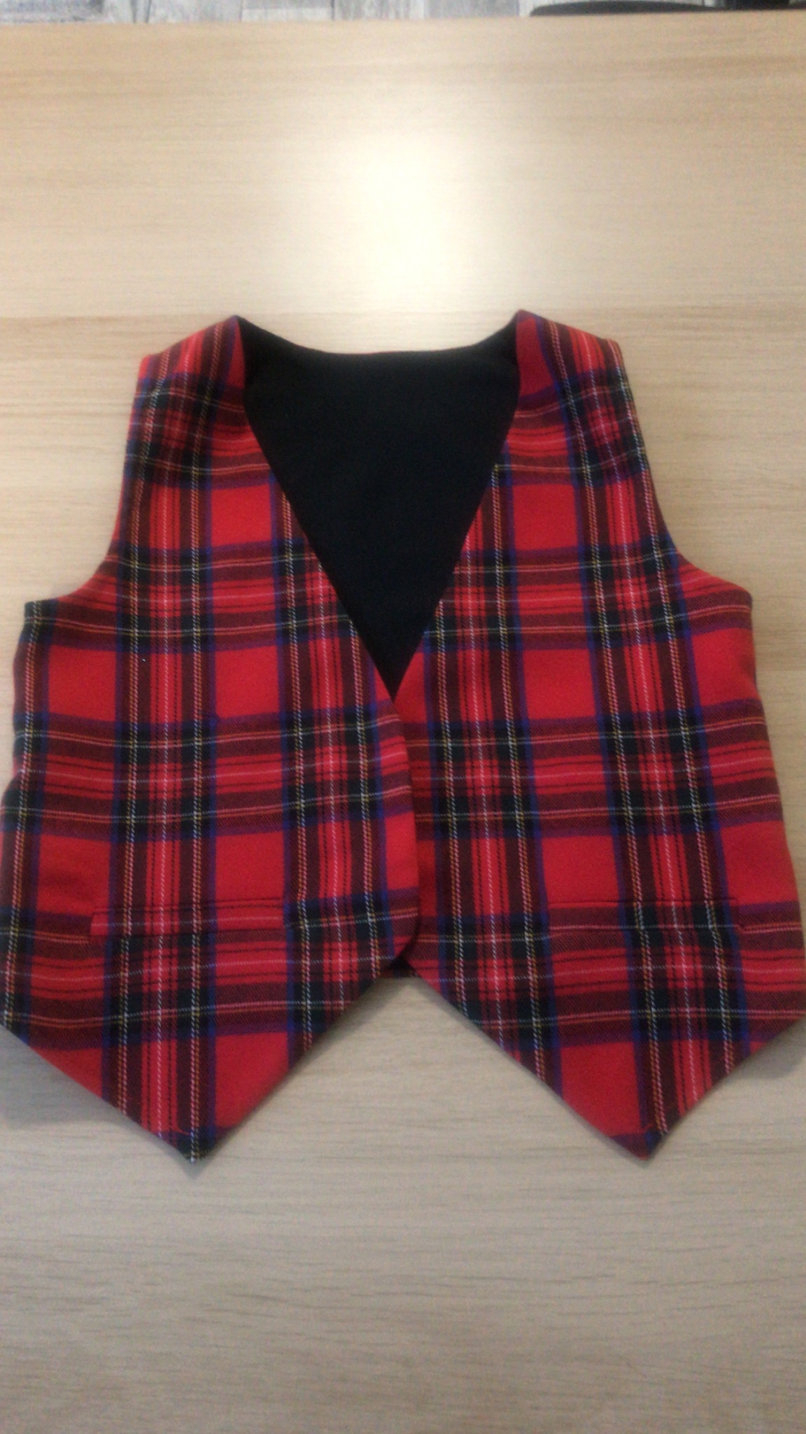 Czerwona kamizelka w kratę szkocką z czarną podszewką, leżąca płasko na drewnianym blacie.