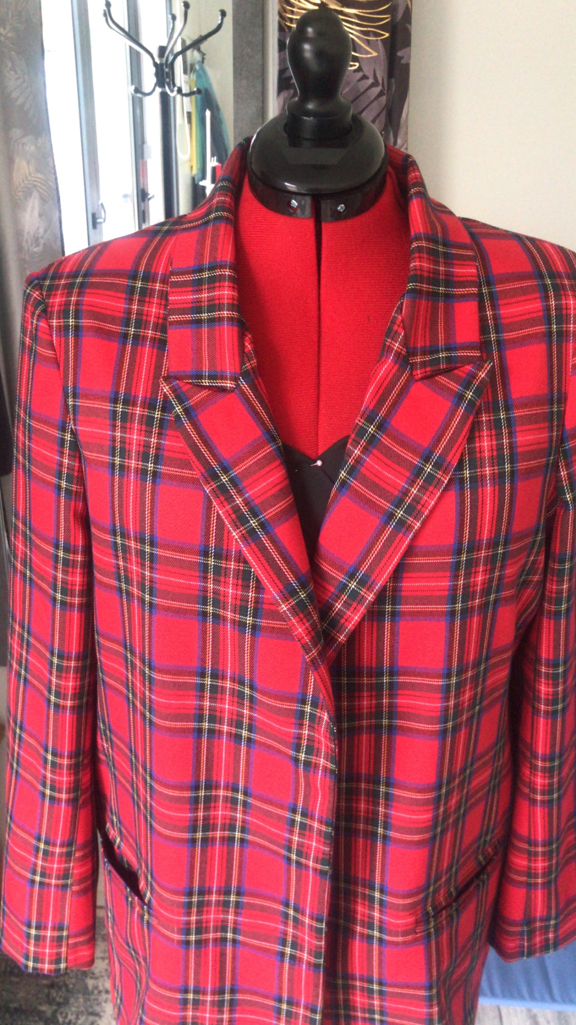 Czerwony, kraciasty żakiet na manekinie krawieckim, widoczne klapy i kieszenie, w tle zasłony i wieszak.
