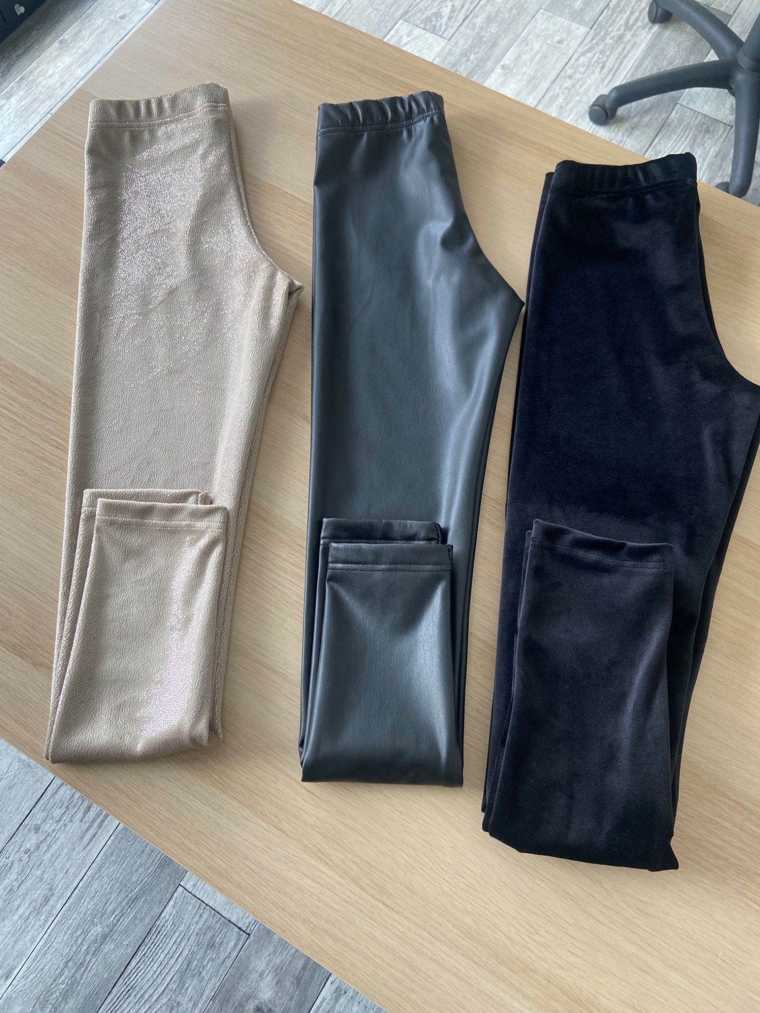 Trzy pary damskich legginsów: beżowe z połyskiem, czarne imitujące skórę i czarne welurowe, ułożone obok siebie na drewnianym blacie.