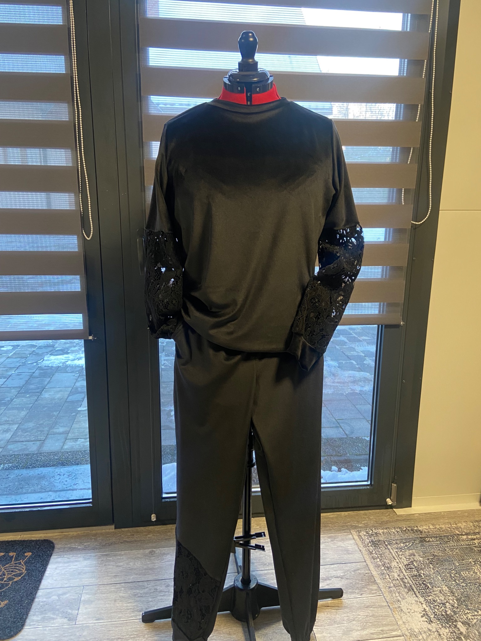 Czarny, dwuczęściowy damski komplet z koronkowymi wstawkami na rękawach i nogawkach, zaprezentowany na manekinie krawieckim na tle okna z żaluzjami.