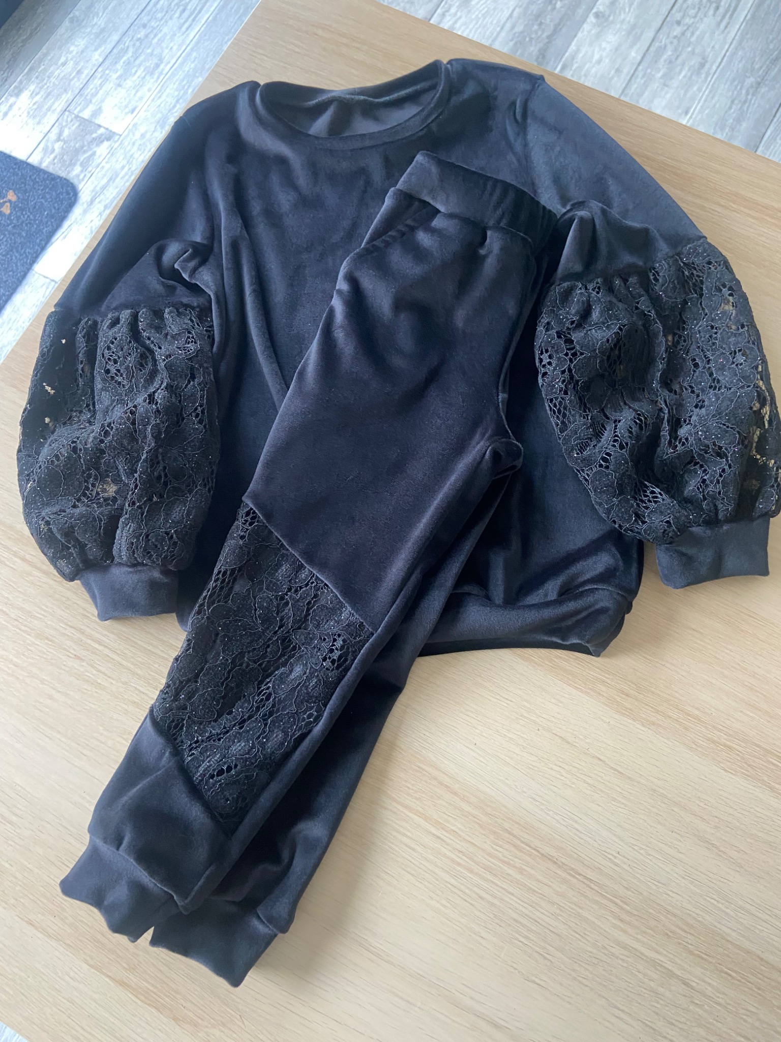 Czarny welurowy komplet dresowy z koronkowymi wstawkami na rękawach bluzy i nogawkach spodni, leżący na drewnianym stole.
