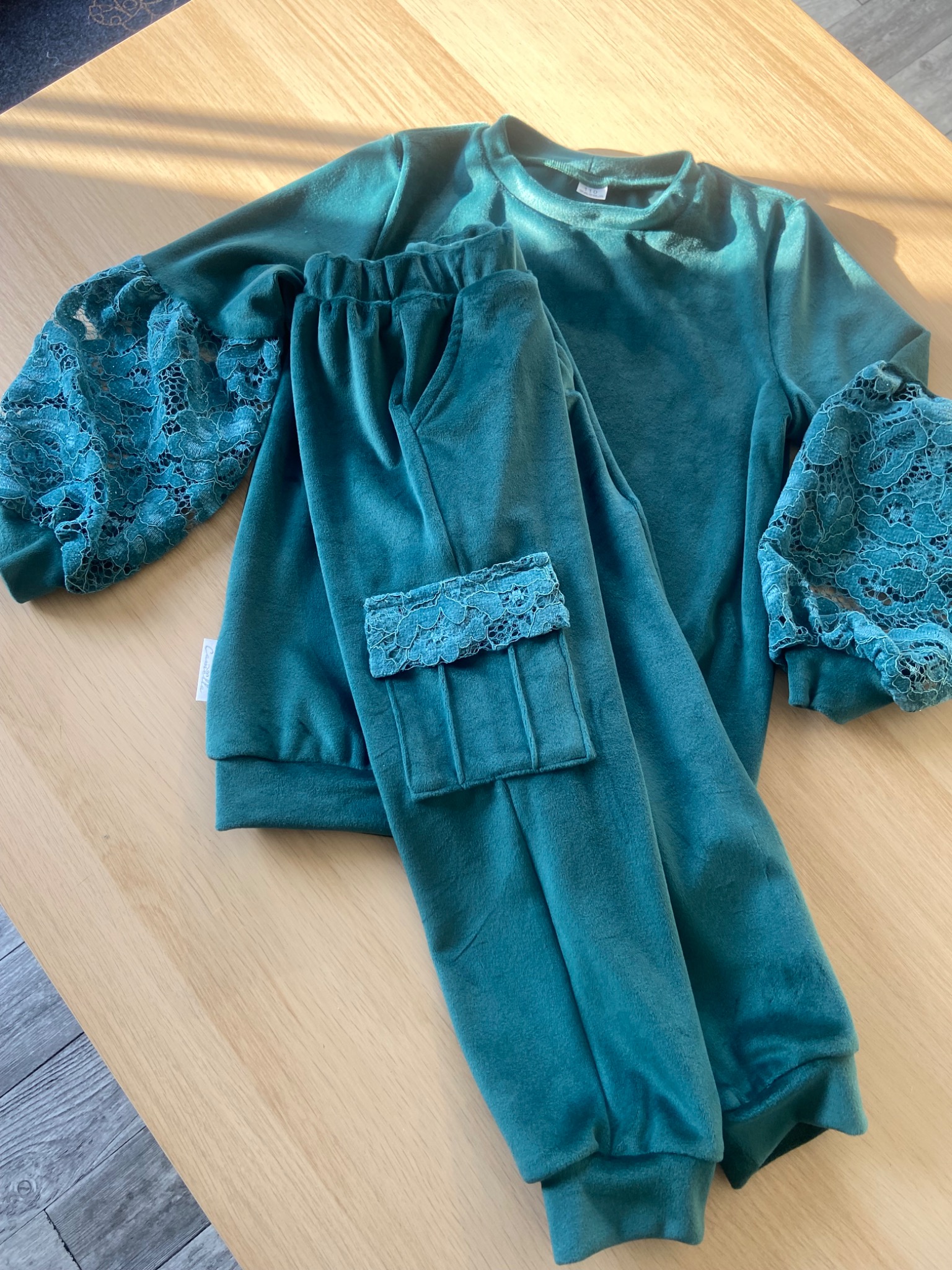 Szmaragdowy welurowy dres dziecięcy z koronkowymi wstawkami na rękawach i kieszeni spodni, leżący na drewnianym stole w słonecznym świetle.