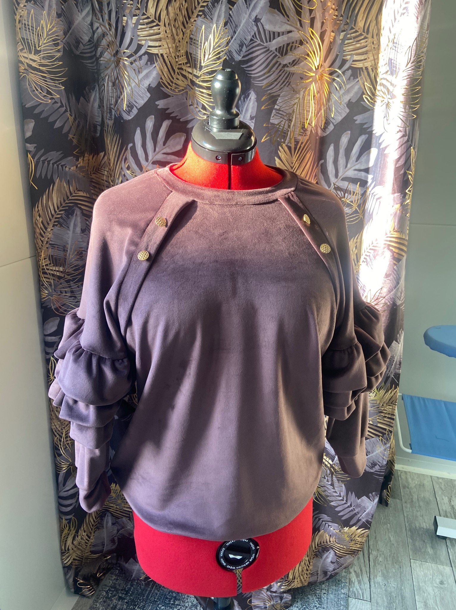 Fioletowa bluza z falbanami na manekinie krawieckim, z detalami w postaci złotych guzików, na tle zasłony w liście.