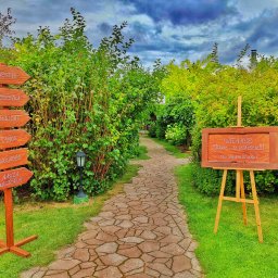 Karczma Maciejówka - Ogród z kamienną ścieżką prowadzącą do tablicy powitalnej na sztaludze oraz drewnianego drogowskazu z napisem 'Wesele', 'Wódeczka', 'Chillout', 'Tańce', 'Wolność', 'Nasza Historia' na tle zieleni...